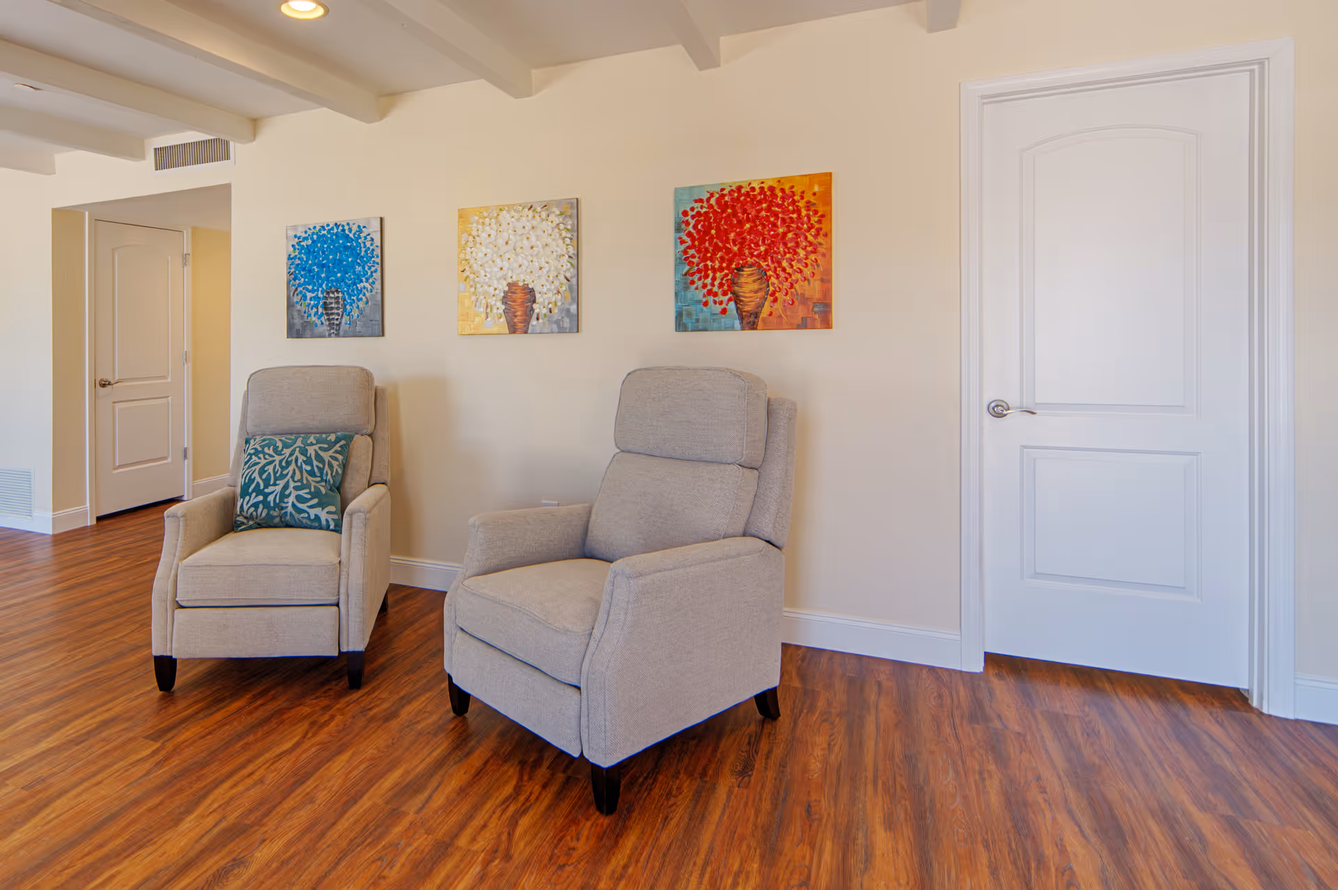A cozy interior room with two beige upholstered armchairs on a wooden floor. One chair has a decorative blue pillow with a coral pattern. Three colorful paintings of vases with flowers in blue, white, and red hang on the wall behind the chairs. There are two white doors visible in the room.