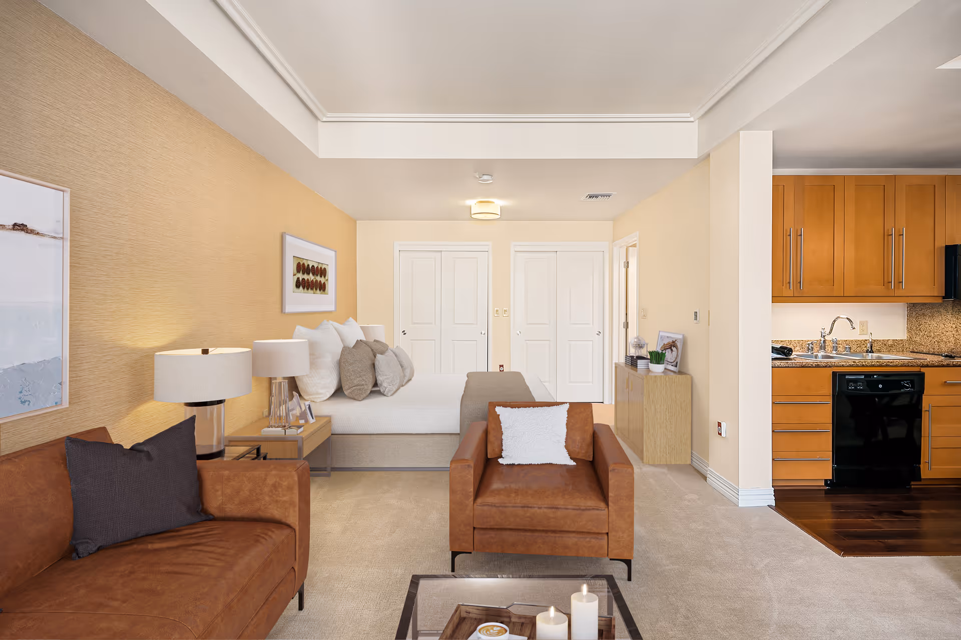 A modern studio apartment with a brown leather sofa and armchair, a bed with multiple pillows, two bedside tables with lamps, a small wooden cabinet with decor items, and a kitchen area with wooden cabinets, a sink, and a dishwasher. The walls are painted in neutral tones and there is framed artwork on the walls.