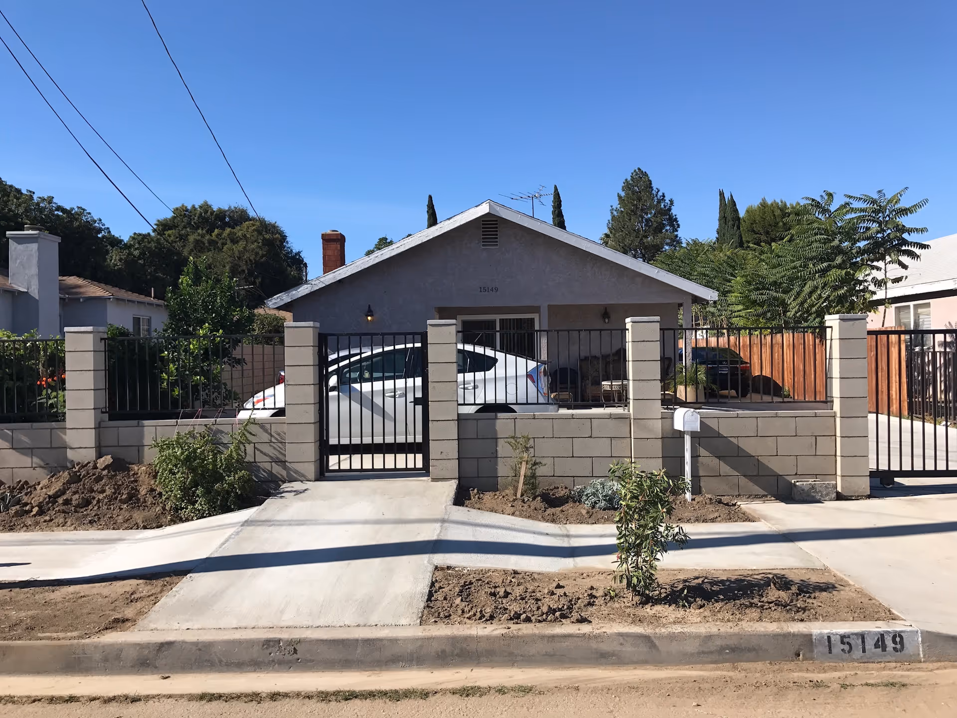 Front exterior view of a single-story house with a gray facade, a chimney, and a gated front yard with a concrete pathway leading to the entrance. A white car is parked inside the gated area, and there are trees and shrubs around the property under a clear blue sky.