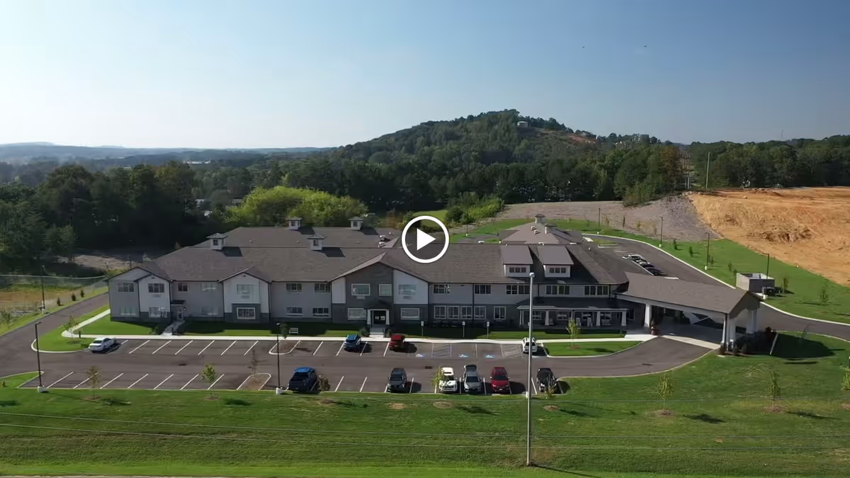 Aerial view of a large, two-story senior living facility building with a parking lot in front, surrounded by green lawns and trees, with hills in the background under a clear sky.