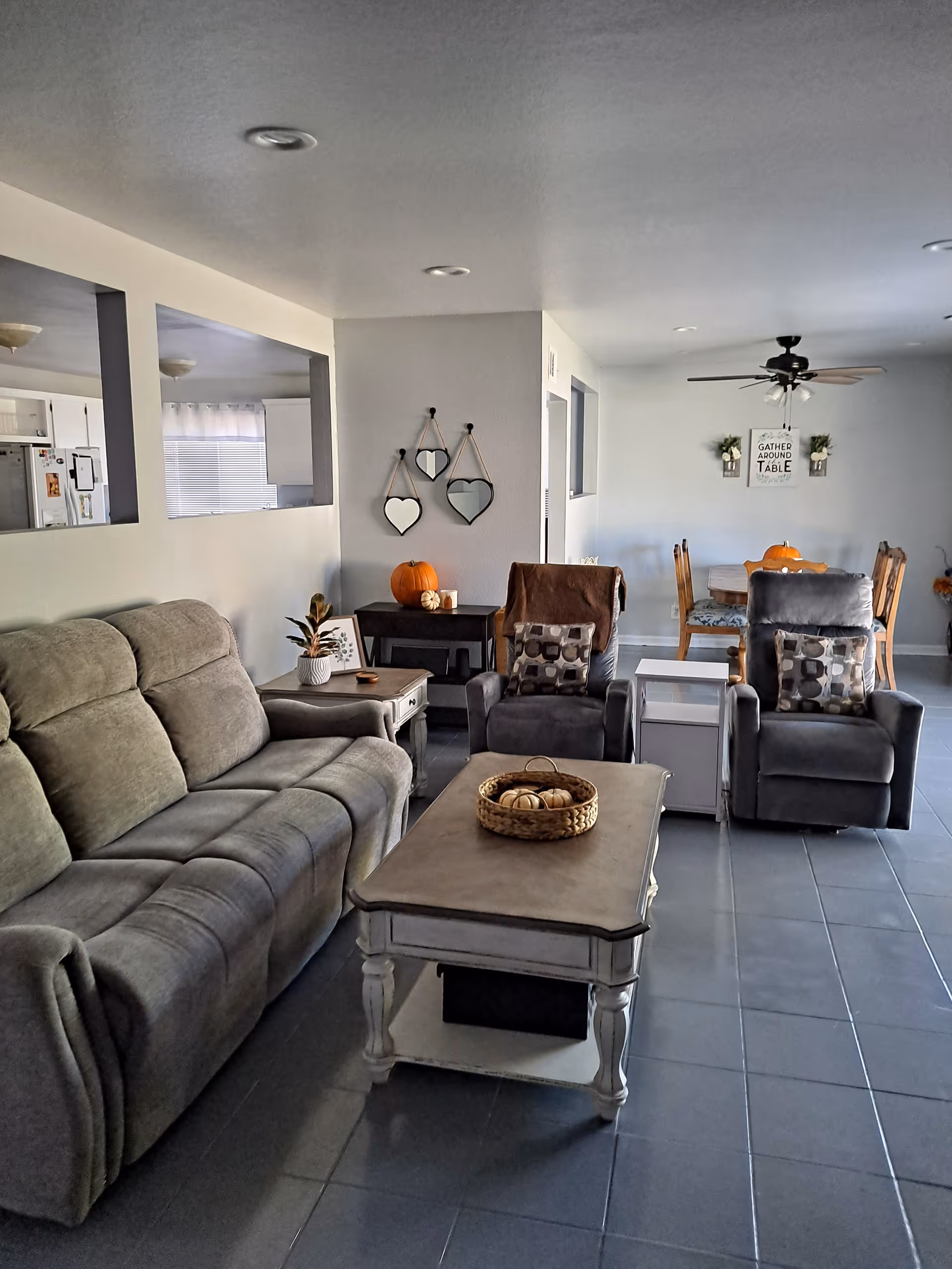 A cozy senior living room with a gray sofa, two gray armchairs with patterned cushions, a white coffee table with a basket of decorative pumpkins, and a side table with a plant and a pumpkin. The room opens into a dining area with a wooden table and chairs, a ceiling fan, and a wall decoration that reads 'Gather Around the Table'.
