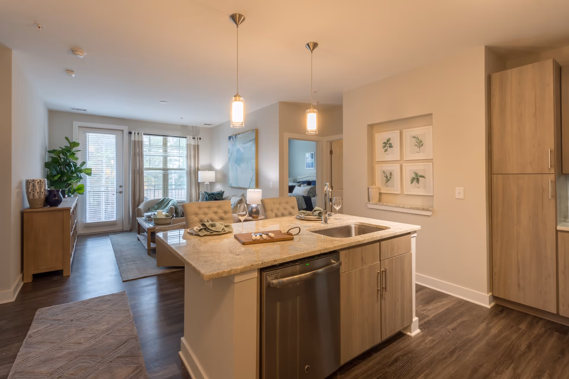 Modern open-concept apartment interior featuring a kitchen island with a sink and two pendant lights above. The island has two cushioned chairs and is set with plates, napkins, and wine glasses. In the background, there is a living area with a sofa, coffee table, large window with curtains, and a door leading outside. Light wood cabinetry and flooring complement the neutral wall colors, with framed botanical artwork on the wall.