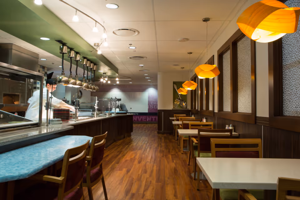 Interior view of a dining area in a senior living facility with a serving counter on the left where a chef is preparing food. There are several tables and chairs arranged along the right side under warm yellow hanging lamps, with wooden flooring and decorative wall panels.
