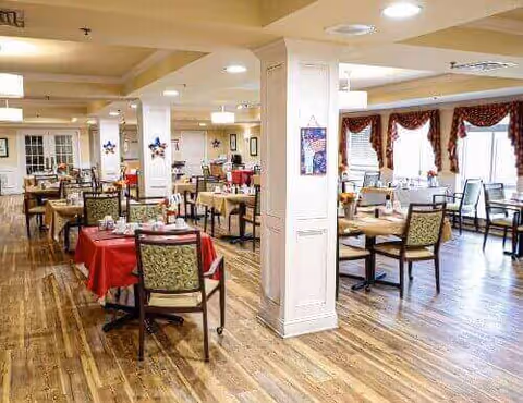 A spacious dining room with wooden floors and multiple tables covered with red and beige tablecloths. Each table is set with plates, glasses, and napkins. The room has large windows with red curtains, white pillars, and decorative wall hangings, creating a warm and inviting atmosphere.