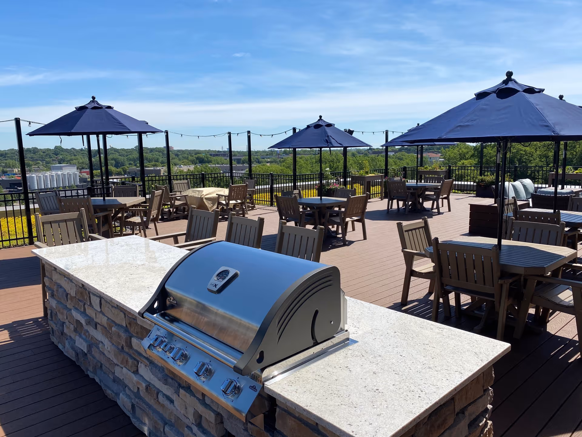 Rooftop terrace with a built-in stainless steel grill, multiple tables and chairs under navy umbrellas, and a tree-lined skyline in the distance.