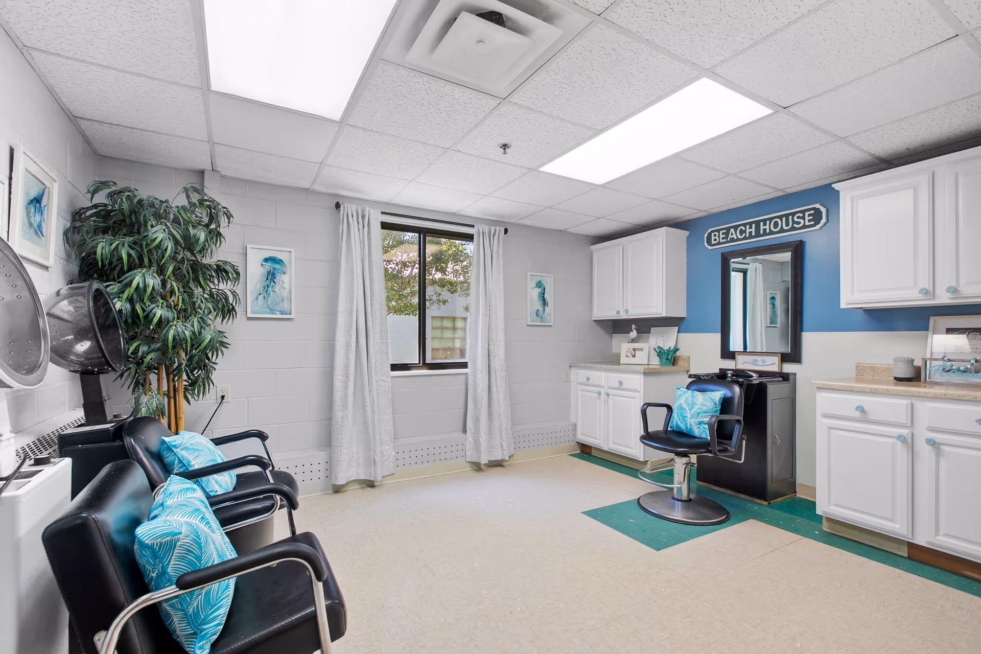 Interior of a bright room with white walls and ceiling tiles, featuring black salon chairs with blue patterned pillows, a large artificial plant, white cabinets, a mirror, and a sign that reads 'BEACH HOUSE'. There are framed pictures of sea creatures on the walls and a window with white curtains letting in natural light.