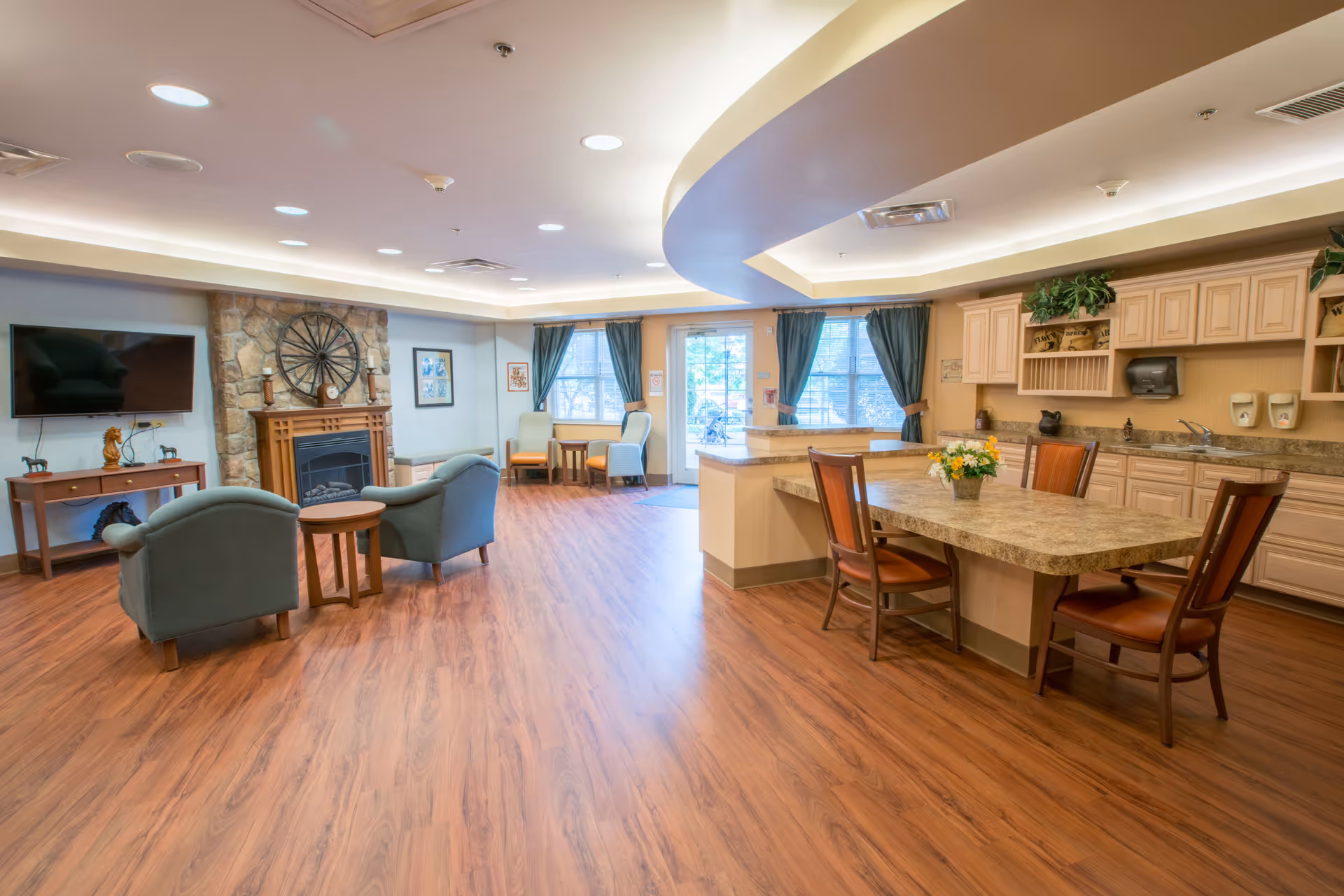 A spacious senior living common area featuring a cozy seating arrangement with two armchairs and a small table in front of a stone fireplace. To the right, there is a kitchen area with a large island countertop surrounded by four chairs, cabinets, and a sink. The room has wood flooring, recessed lighting, and large windows with dark curtains allowing natural light to enter.