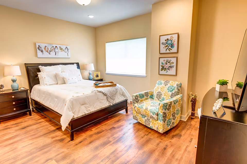 A cozy bedroom with a wooden bed frame and white bedding, two bedside tables with lamps, a window with blinds, a floral patterned armchair, two framed floral artworks on the wall, and a dark wooden dresser with a TV and small decorative plants.