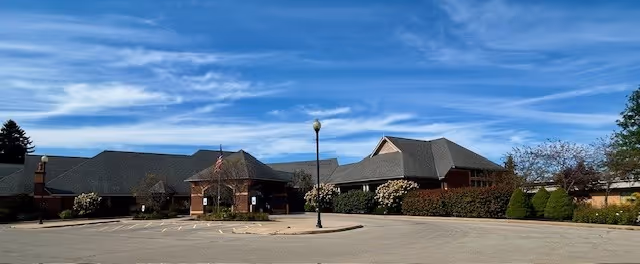 Exterior view of Orchard Manor facility showing a large brick building with multiple roof peaks under a blue sky with scattered clouds. The foreground includes a paved parking area with a single lamppost and some landscaped bushes and trees around the building.