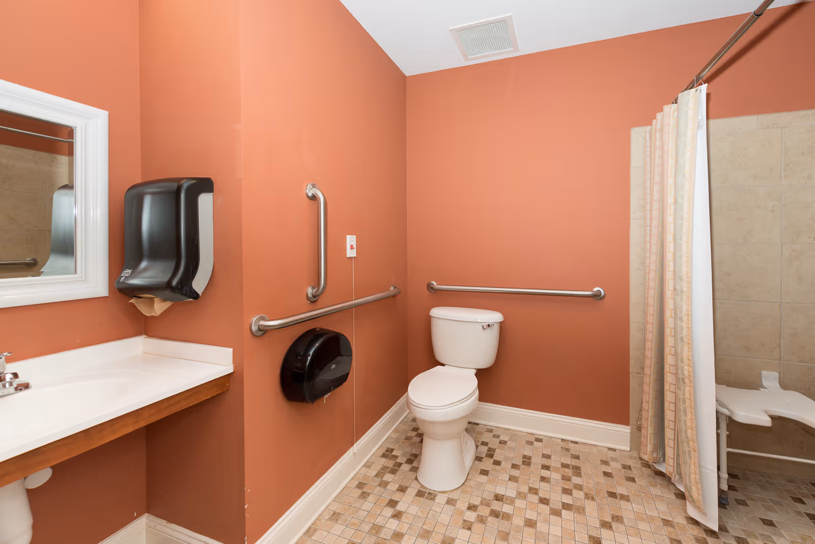 Accessible bathroom with a toilet, grab bars, sink and paper dispensers, and a curtained shower area.
