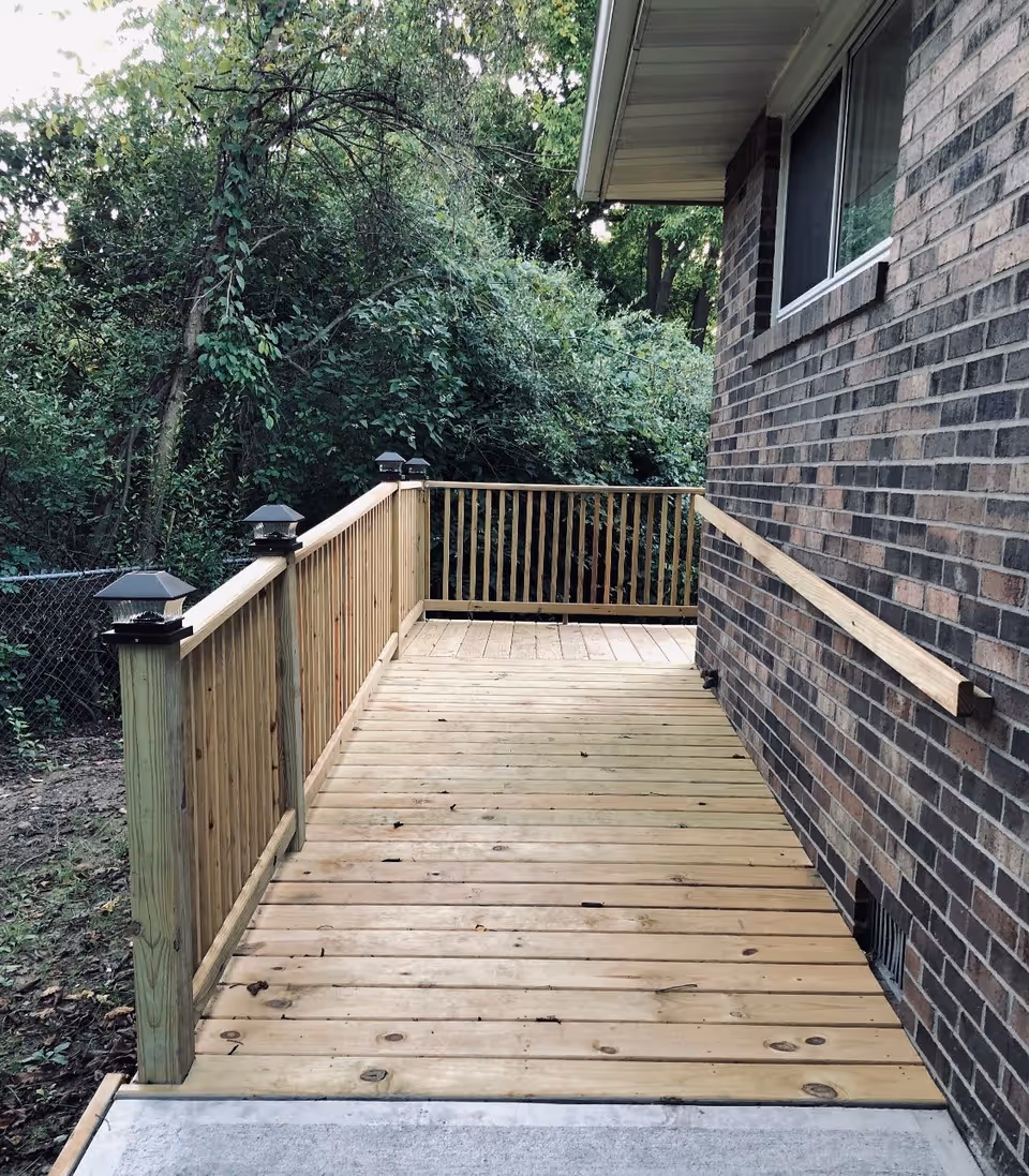 Wooden exterior deck with railings and a wall-mounted handrail alongside a brick house overlooking a wooded yard.