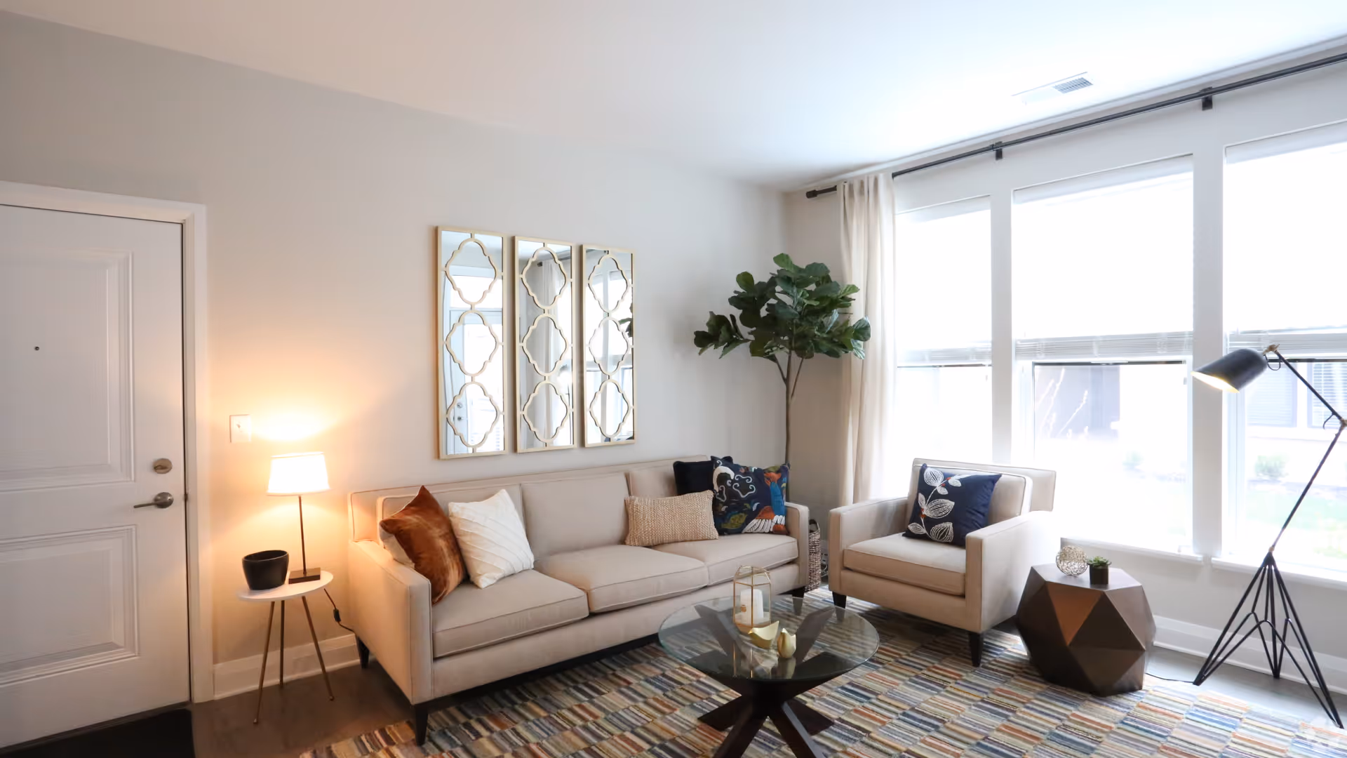 A bright living room with a beige sofa adorned with multiple decorative pillows, a matching armchair with a patterned pillow, a round glass coffee table with decorative items, a geometric side table with small plants, a floor lamp, a potted plant in the corner, three decorative mirrors on the wall, and large windows with white curtains allowing natural light to fill the room.