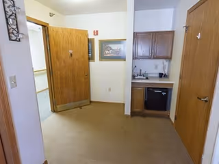 Interior view of a small room in Northern Oaks Place showing an open wooden door, a small kitchenette area with a sink, countertop, cabinets, and a mini refrigerator, beige carpeted floor, and a framed picture on the wall.