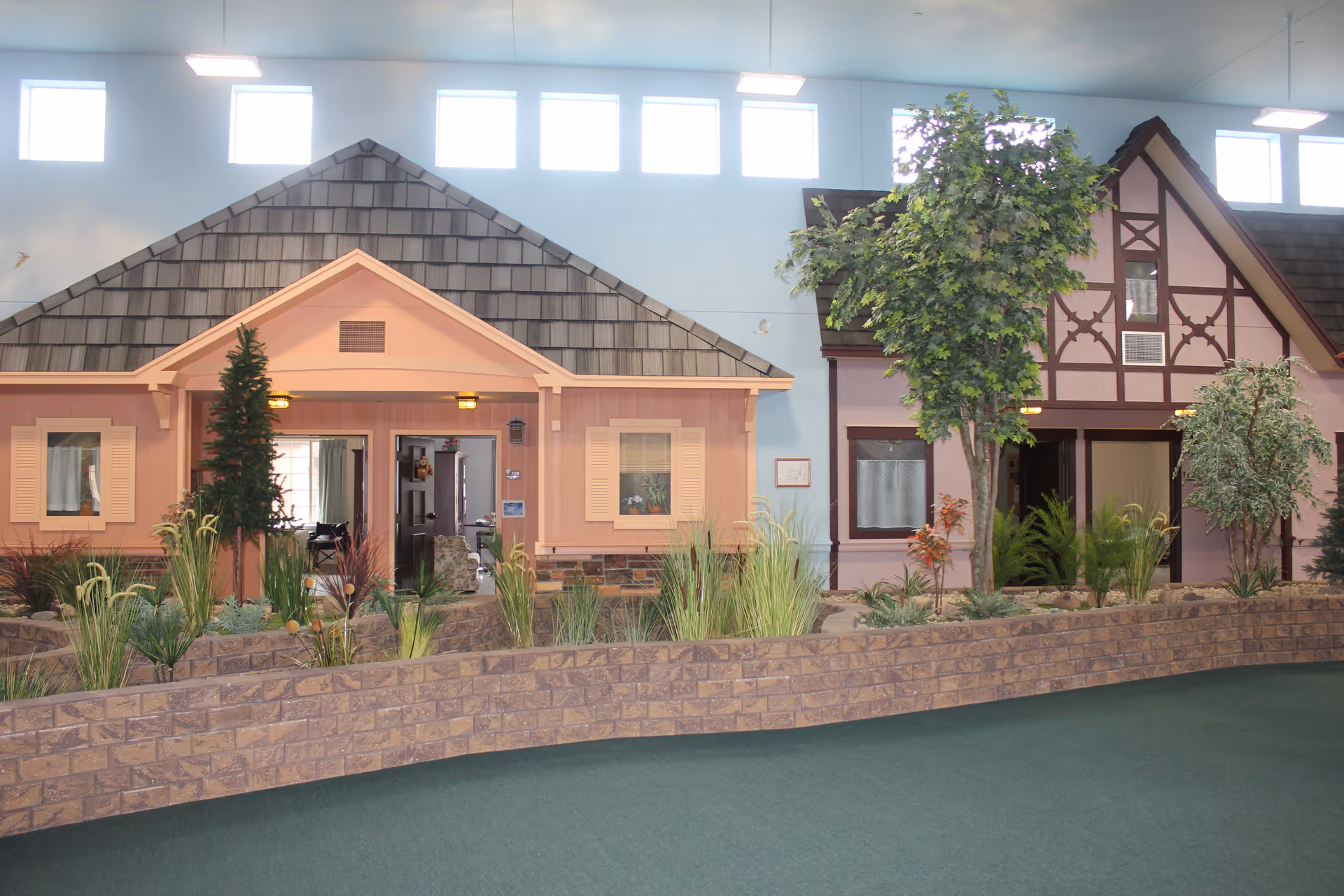 Indoor scene showing two house facades with pitched roofs and windows, surrounded by artificial plants and trees, with a blue wall and high windows in the background.