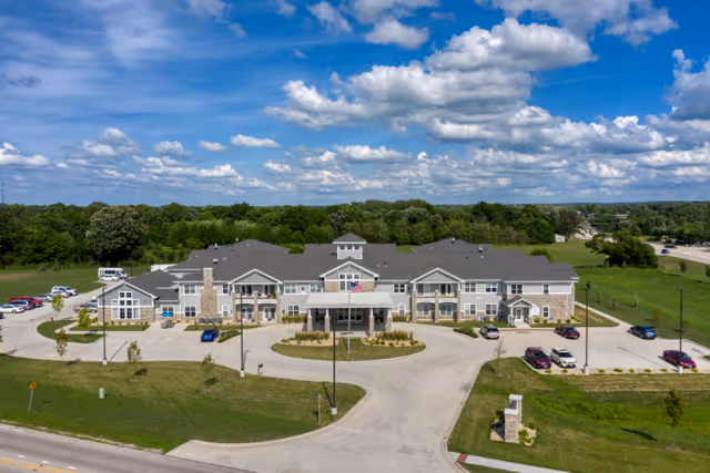 Aerial front view of a two-story senior living facility with a circular driveway, flagpole, and surrounding lawn under a blue sky.