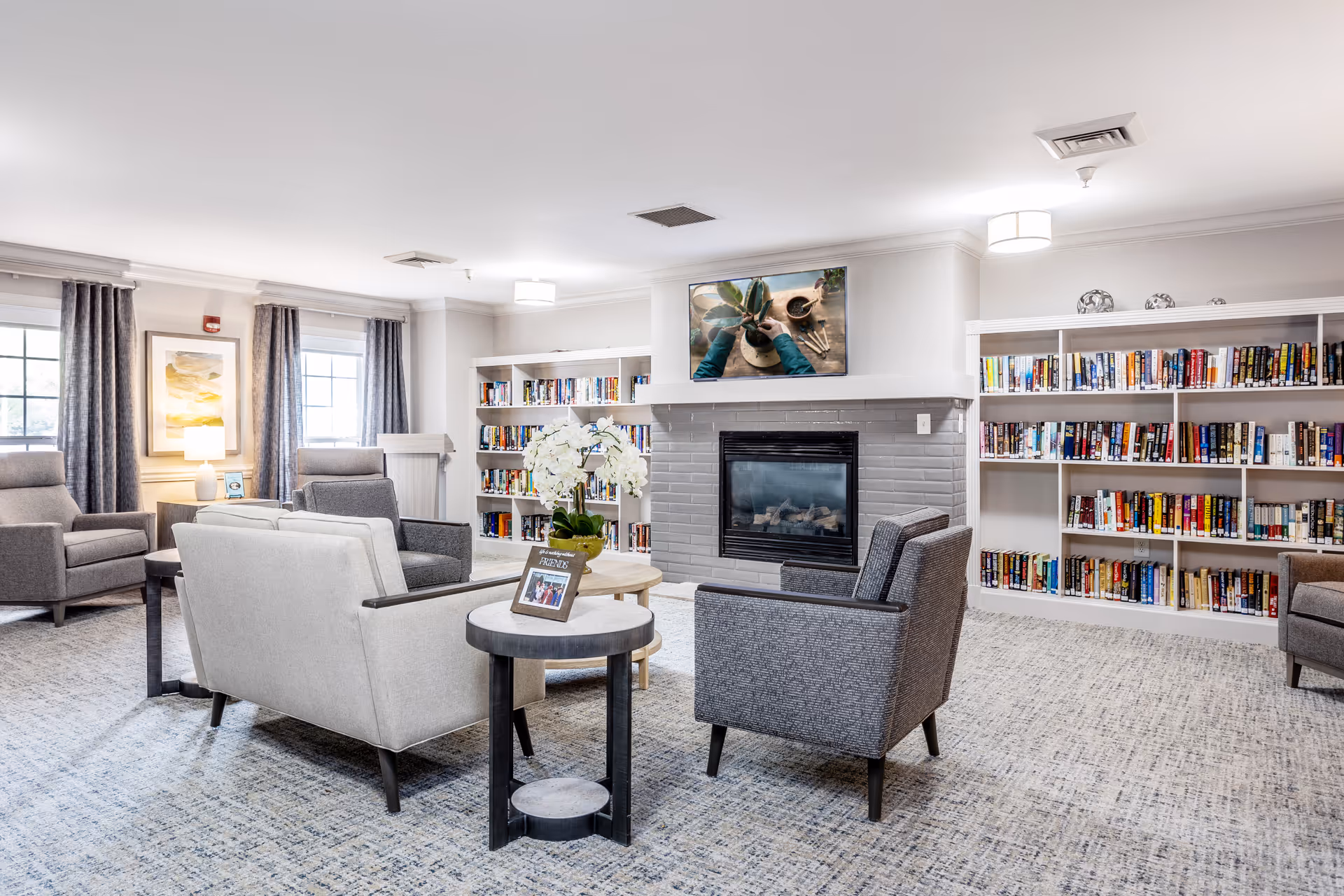 A bright and cozy living room area with multiple armchairs arranged around a small round table with a flower vase. There is a fireplace with a TV mounted above it, and bookshelves filled with books line the walls. Large windows with curtains allow natural light to fill the room.