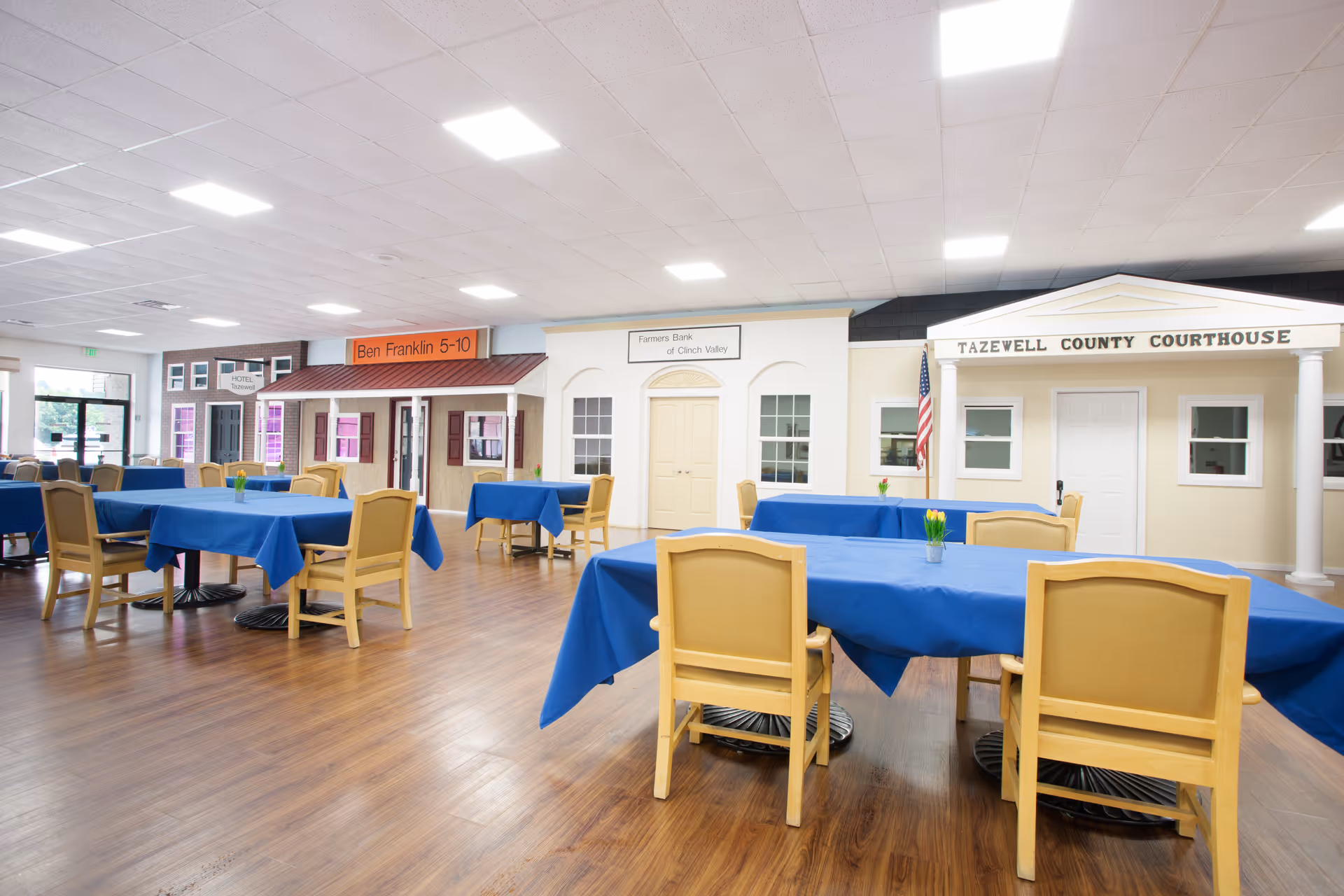 Large bright dining room with blue-tablecloth-covered tables and yellow chairs on wood floors in front of decorative storefront facades.