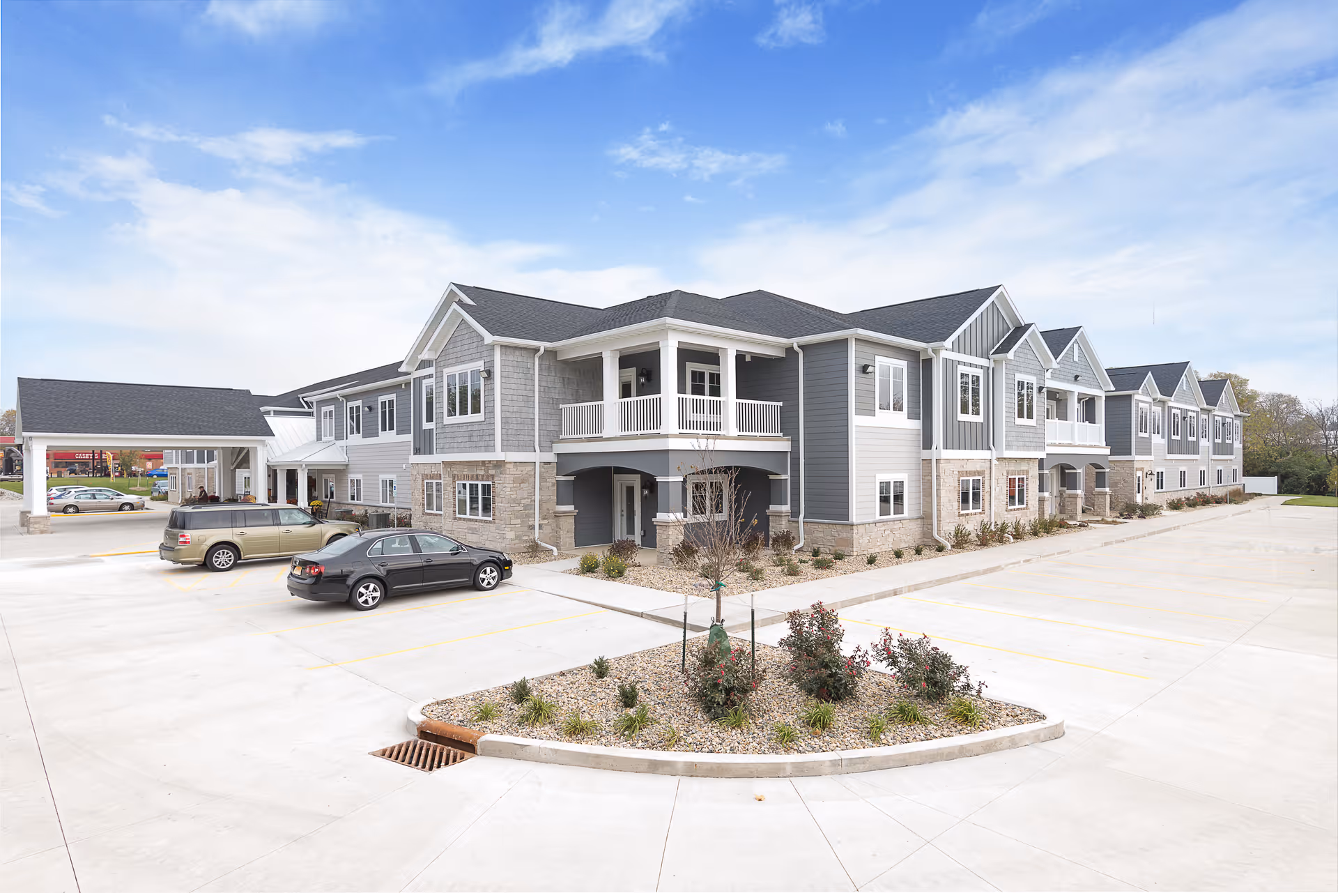 Two-story gray-and-white assisted living building with balconies, a landscaped island, and parked cars in a large front parking area under a blue sky.
