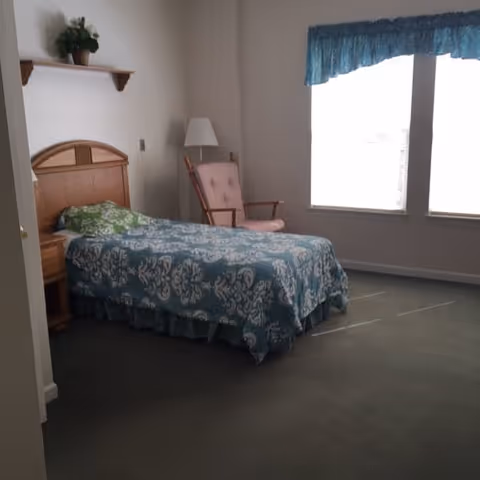 A simple bedroom with a single bed covered in a blue and white patterned bedspread, a wooden headboard, a small wooden nightstand, a pink cushioned rocking chair, a floor lamp, and two large windows with blue valances letting in natural light.
