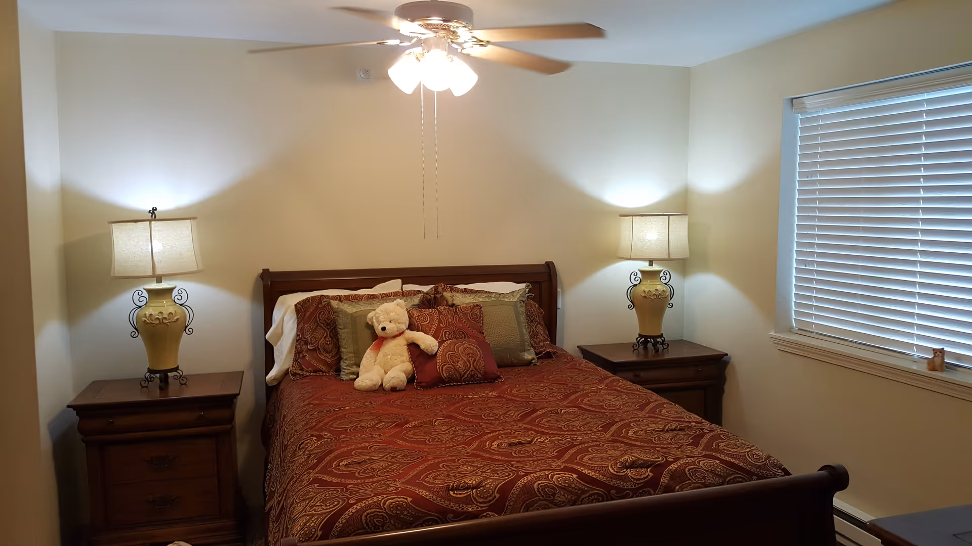 Cozy bedroom with a made bed topped by a teddy bear, matching bedside tables and lamps, a ceiling fan, and a window with blinds.