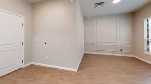 Empty interior room with wood-look floors, decorative wall paneling, recessed ceiling lights and a window with blinds.