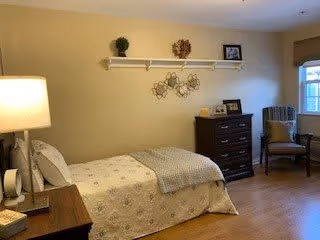 A cozy bedroom with a single bed covered in a light-colored patterned bedspread. Next to the bed is a wooden nightstand with a lamp and a small clock. On the wall above the bed is a white shelf holding decorative items including a small plant, a wreath, and a framed picture. Across from the bed is a dark wooden dresser with framed photos and decorative pieces on top. A cushioned chair is placed near a window with blinds, and the room has wooden flooring and soft yellow walls.