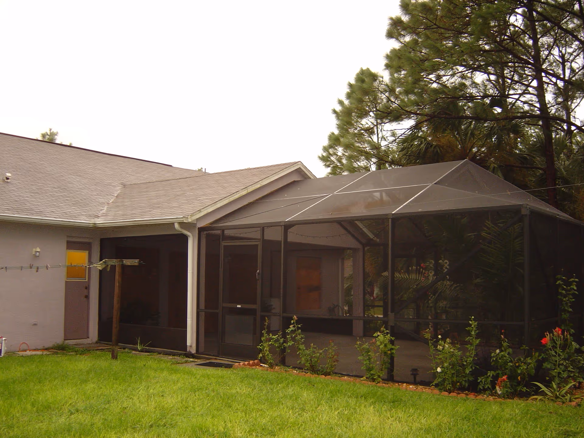 Exterior view of a single-story building with a screened-in patio area surrounded by a garden with green grass and plants. The building has a sloped roof and a door with a yellow window on the left side.