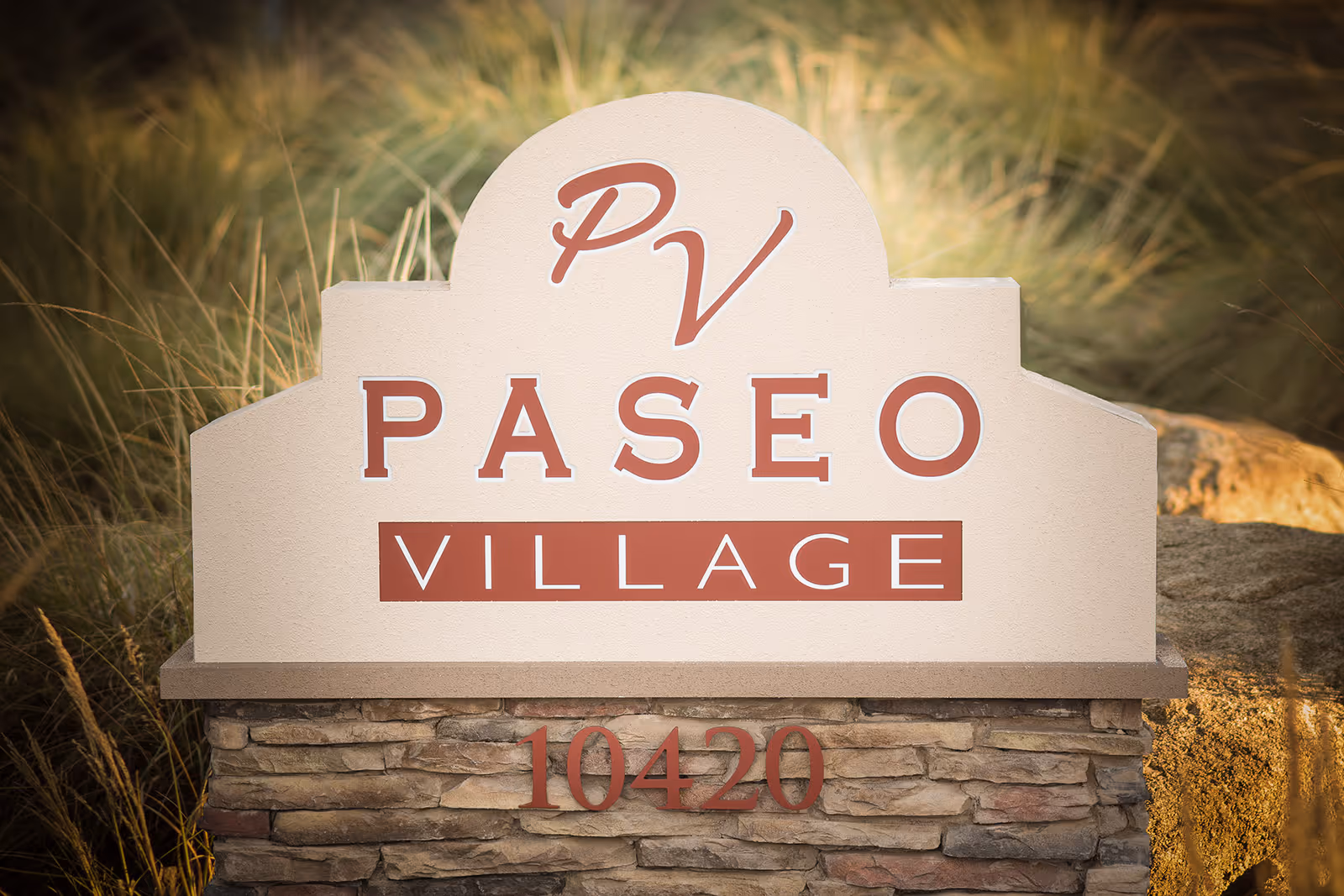Stone and stucco sign for Paseo Village with the letters PV at the top and the number 10420 below, surrounded by tall grass and rocks.