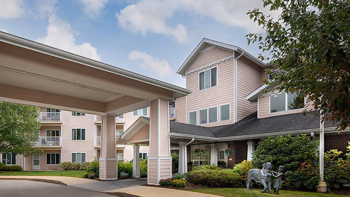 Exterior view of Holiday Maple Suites senior living facility showing a covered entrance with columns, multiple windows, and a landscaped garden area with a statue of a child and a dog wearing sunglasses.