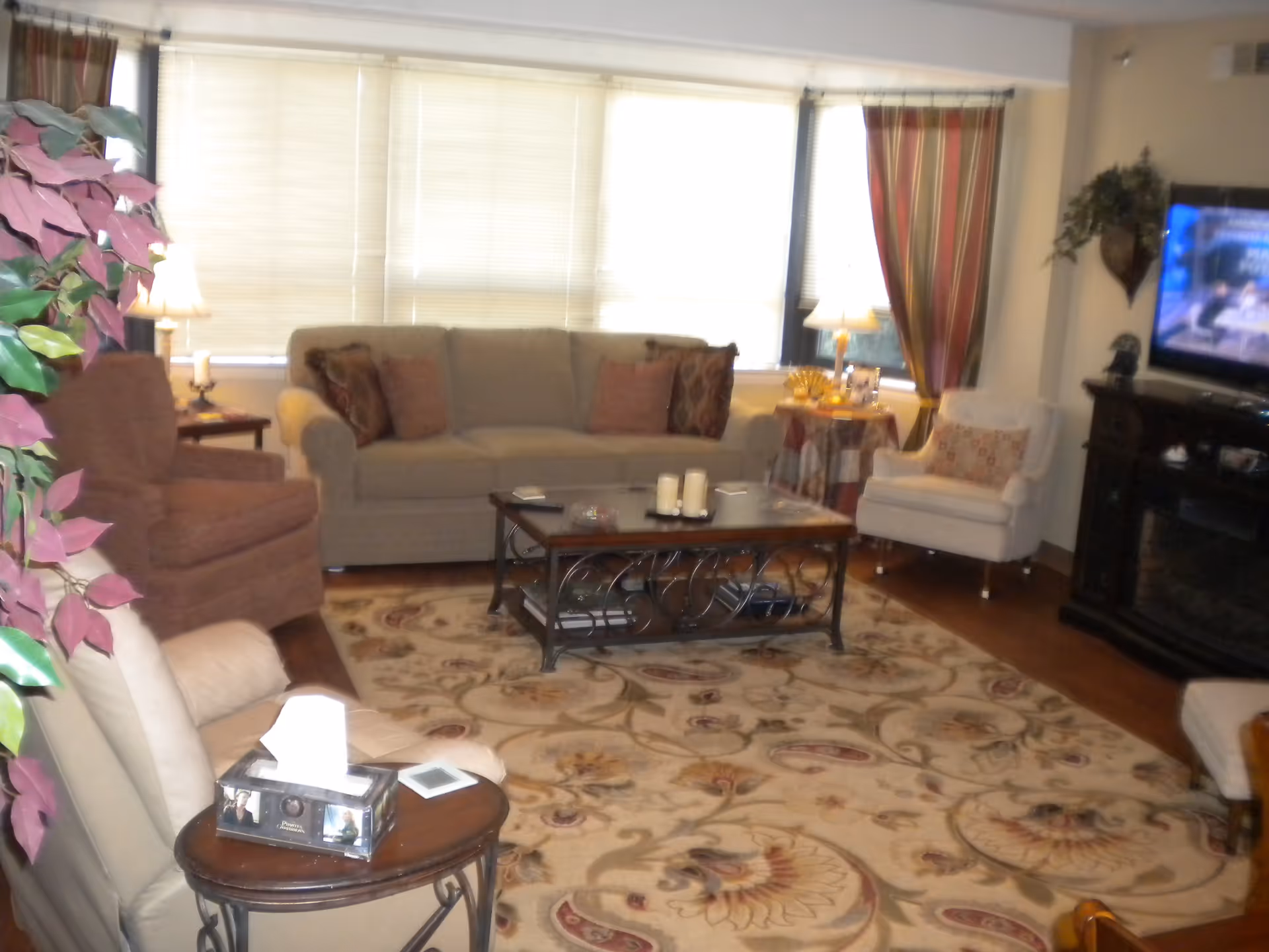 A cozy living room with a beige sofa, two armchairs, a patterned area rug, a wooden coffee table with candles, a side table with a lamp, and a flat-screen TV mounted on a dark wooden stand. There are large windows with blinds and curtains, and a decorative plant is visible on the left side.