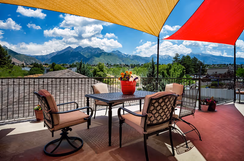 Outdoor patio area with a metal table and four cushioned chairs under yellow and red shade sails, overlooking a scenic view of mountains and greenery under a partly cloudy blue sky. There are potted flowers on the table and around the patio.
