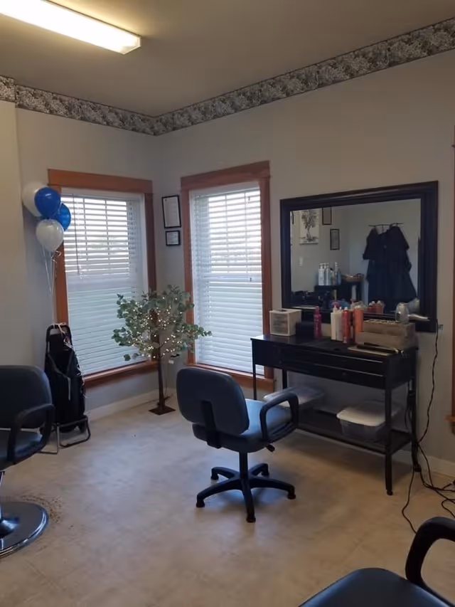 Interior room with two large windows with blinds, a small artificial tree with lights, two salon chairs, and a black vanity table with a large mirror. Various hair care products and tools are on the table. The room has light-colored walls with a floral border near the ceiling and a tiled floor.