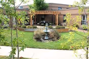 Courtyard with a central fountain, grassy lawn, plants, and a pergola-covered seating area in front of a brick building.