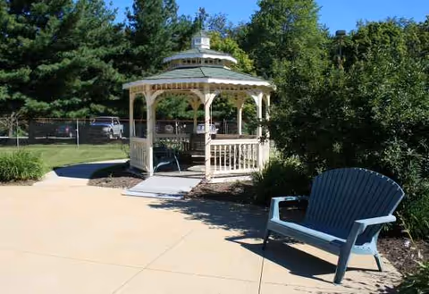 Sunlit gazebo and blue chair sit in a paved outdoor courtyard surrounded by trees.