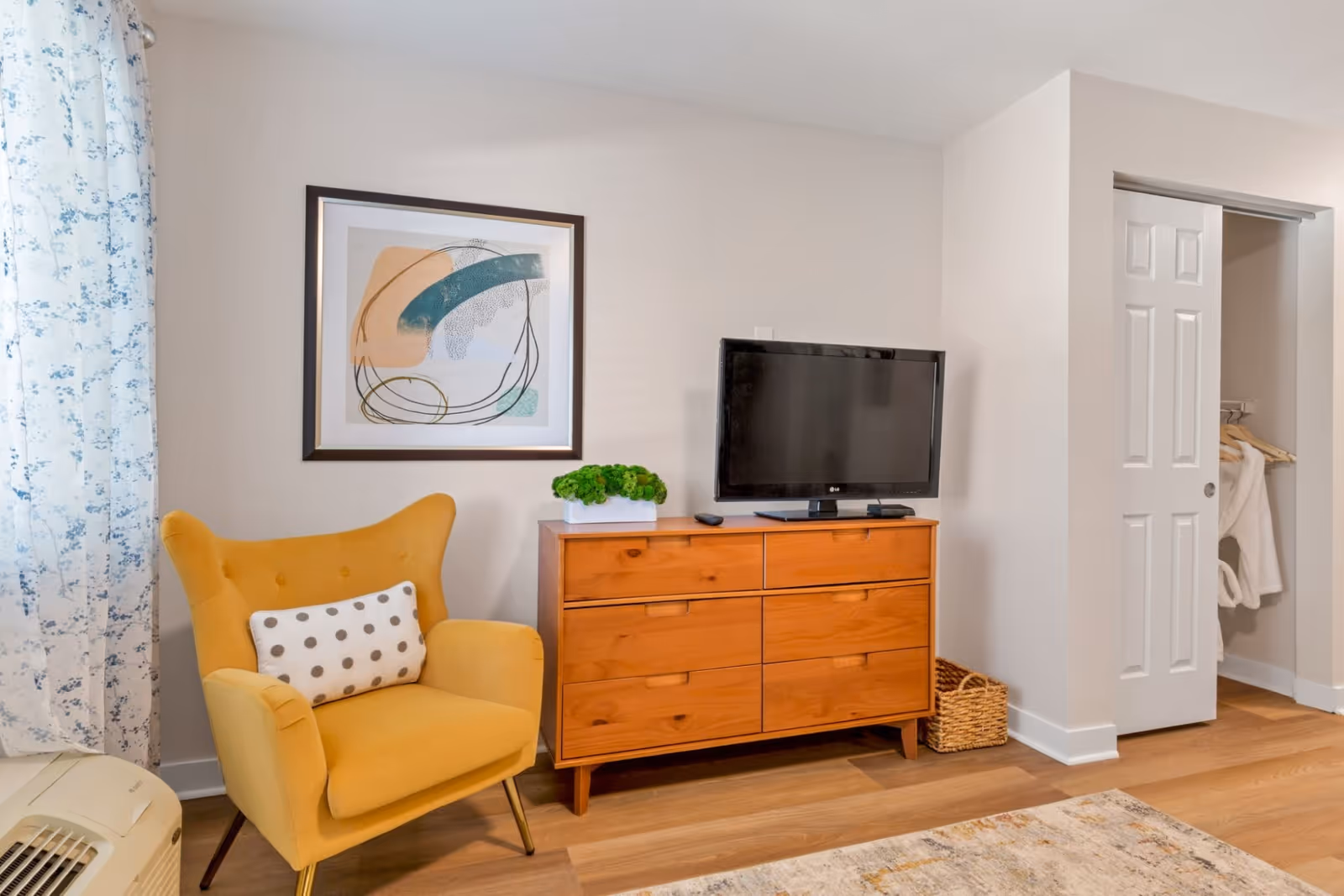 A cozy living space with a mustard yellow armchair featuring a white pillow with gray polka dots, a wooden dresser with six drawers, a flat-screen TV on top, a small green plant, an abstract framed artwork on the wall, a closet with sliding white doors partially open showing hanging clothes, and light wood flooring with a patterned rug.