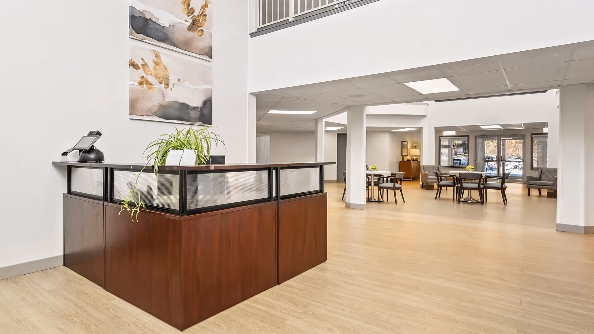 Interior view of a senior living facility lobby with a wooden reception desk featuring a plant and a phone. The space has light wood flooring, white walls, and modern abstract artwork on the wall. In the background, there are tables and chairs arranged in a common area with large windows letting in natural light.