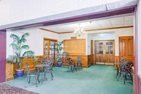 An interior common area with green carpet and light-colored wallpaper. The room contains several black metal tables and chairs with intricate designs. There are two large potted plants with green foliage placed near wooden framed glass doors. A chandelier hangs from the ceiling, and a wooden bulletin board is mounted on the wall. The room has wooden trim and multiple wooden doors.