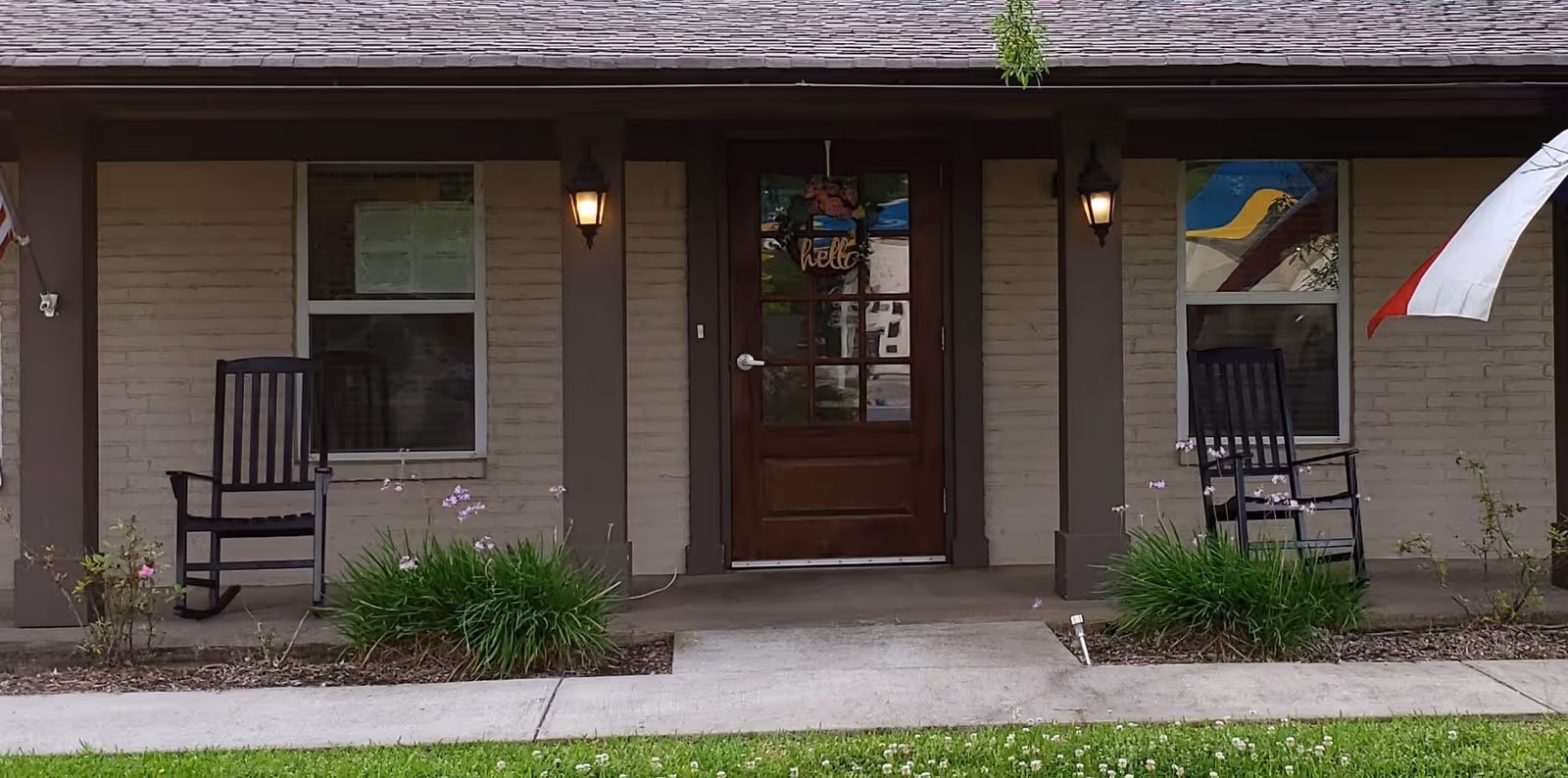 Front porch entrance of a single-story building with a central wooden door flanked by windows, two rocking chairs, porch lights, and small garden beds.