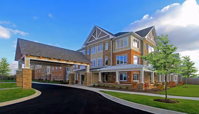 Exterior view of a large, modern three-story senior living facility building with a covered entrance driveway, surrounded by green lawns and trees under a partly cloudy blue sky.