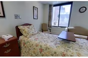 A small bedroom with a single bed covered in floral bedding, a wooden nightstand with a telephone, a chair near a window with curtains, and a wall clock above the window.