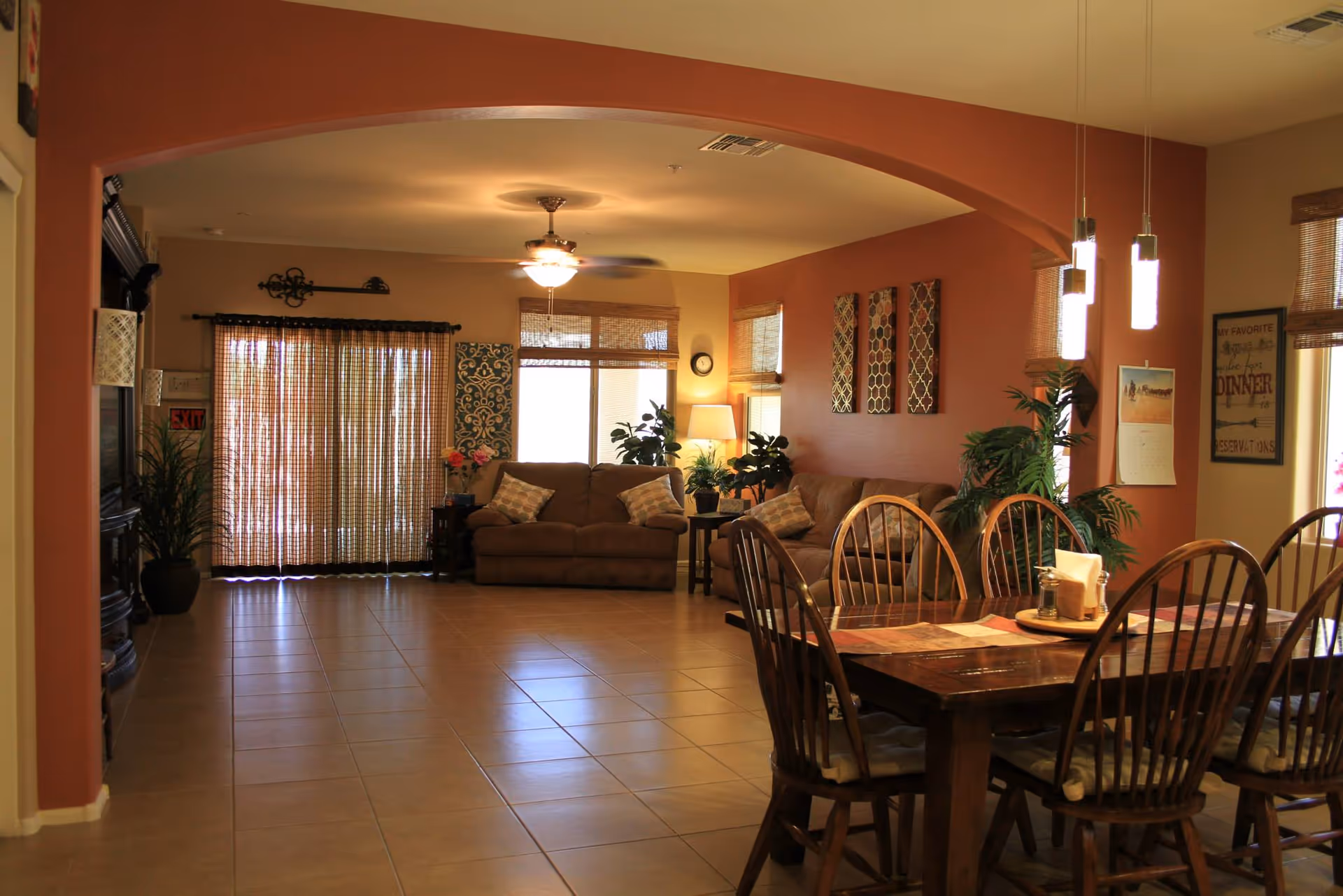 Interior view of a senior living facility showing a combined living and dining area. The living area has two brown sofas with patterned cushions, a floor lamp, wall art, and several potted plants. The dining area features a wooden table with six chairs, pendant lights hanging above, and a window with blinds. The floor is tiled, and the walls are painted in warm tones.
