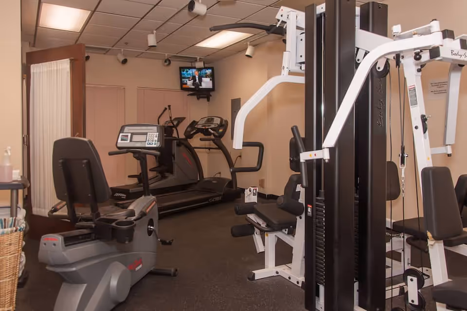 Indoor fitness room with exercise equipment including a recumbent bike, treadmill, elliptical machine, and a multi-station weight machine. A small wall-mounted TV is visible in the background, and the room has beige walls and a tiled ceiling.