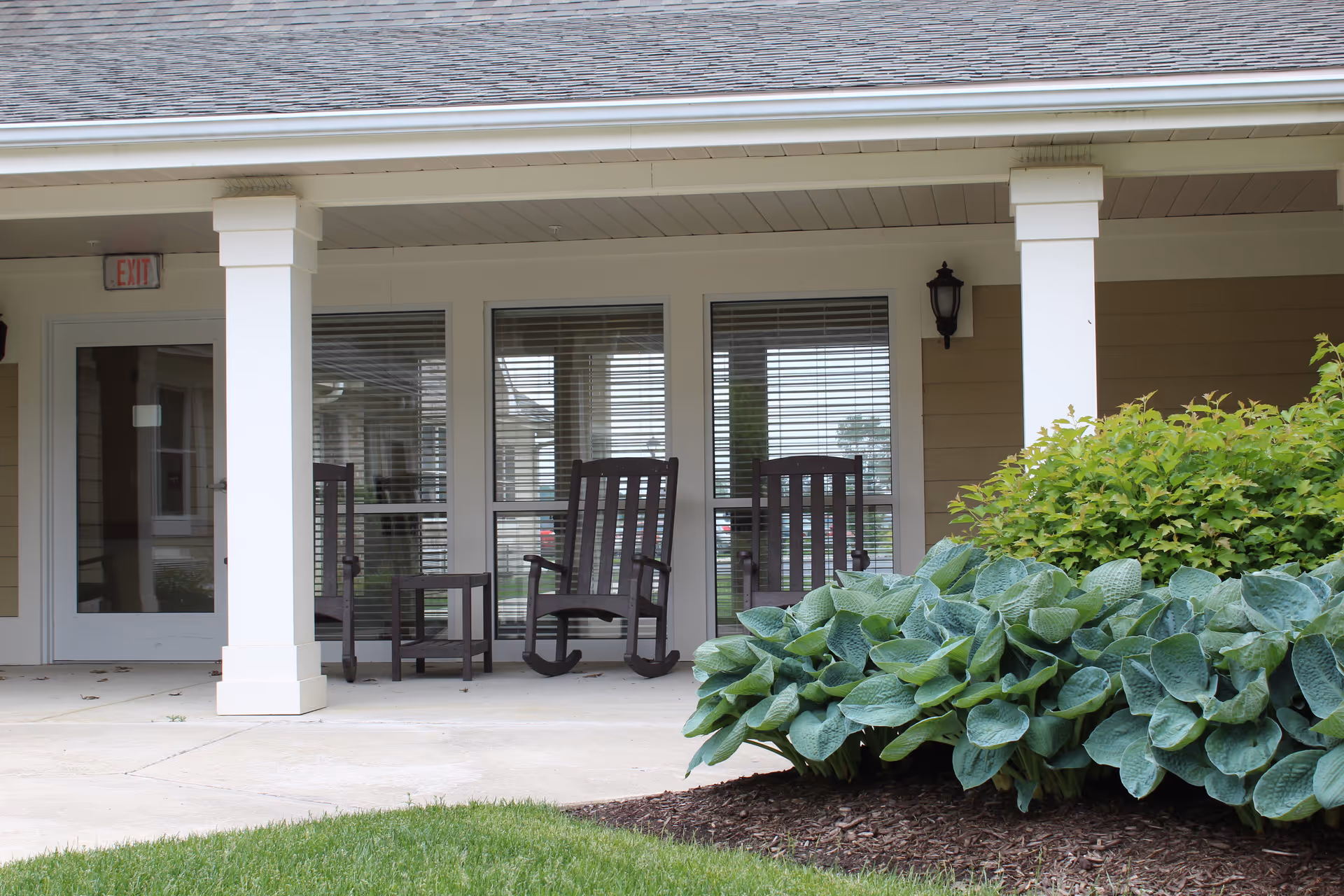 Covered porch area with two wooden rocking chairs and a small table between them, in front of windows with blinds. There are green bushes and plants in the foreground and a concrete walkway leading to the porch.