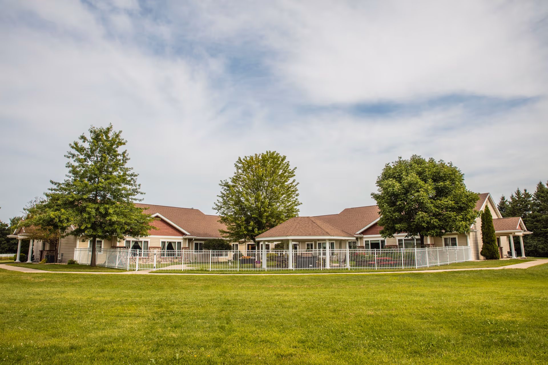 Exterior view of Oakwood Manor, a single-story building with a brown roof and beige siding, surrounded by green grass and several trees under a partly cloudy sky.