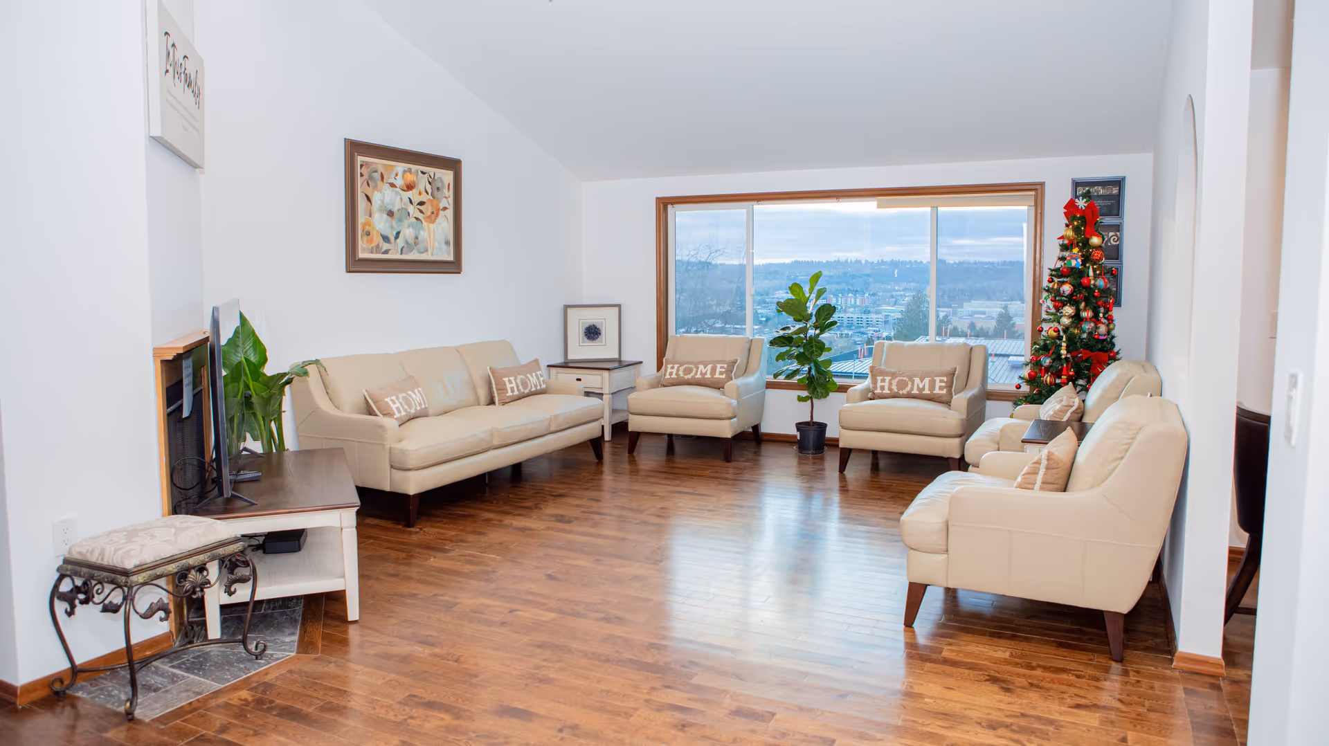 A bright living room with wooden flooring, beige leather sofas and armchairs with 'HOME' pillows, a large window showing an outdoor view, a decorated Christmas tree, a small side table with a plant, and framed artwork on the walls.