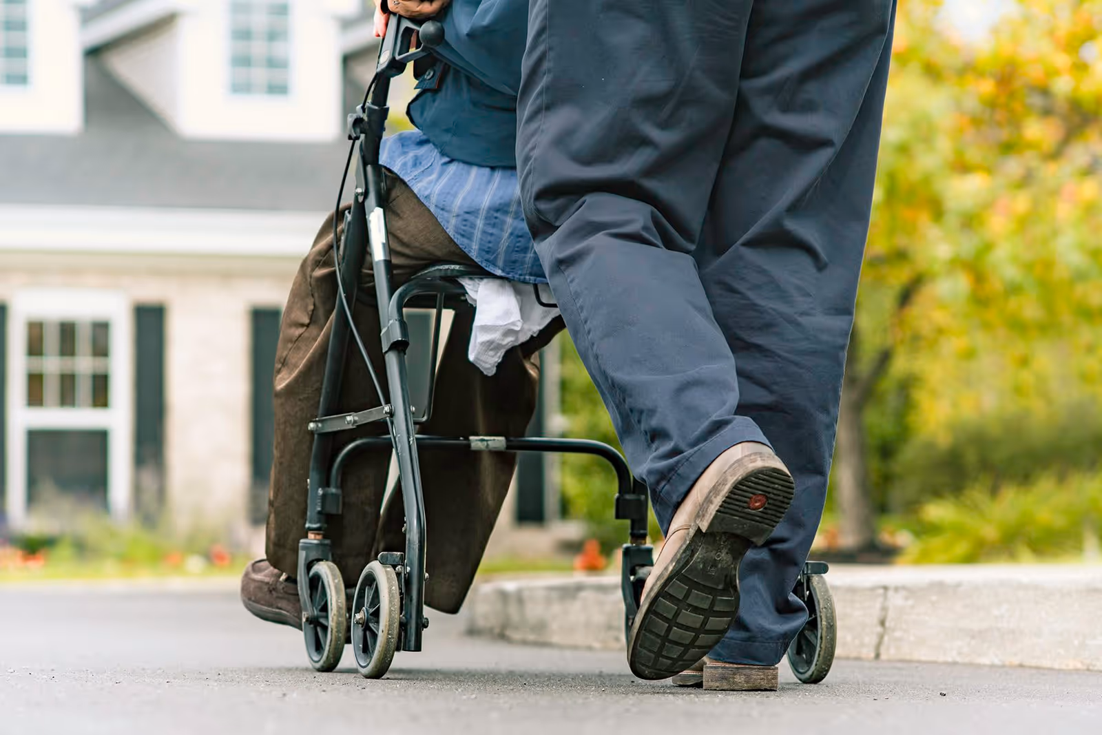 Close-up of an elderly person using a walker being assisted by another person walking beside them outdoors near a residential building.