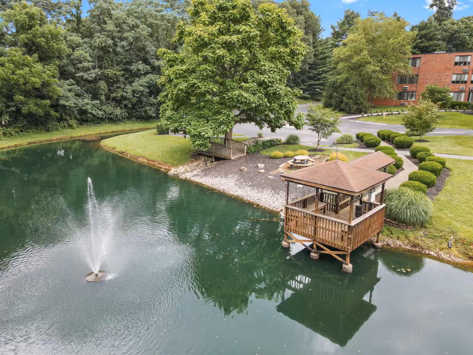 A serene outdoor scene at Brownsboro Park Retirement Community featuring a small pond with a water fountain in the center. There is a wooden gazebo built over the water, surrounded by well-maintained landscaping including bushes, trees, and a grassy area. A picnic table and a small bridge are visible near the pond, with a brick building in the background.