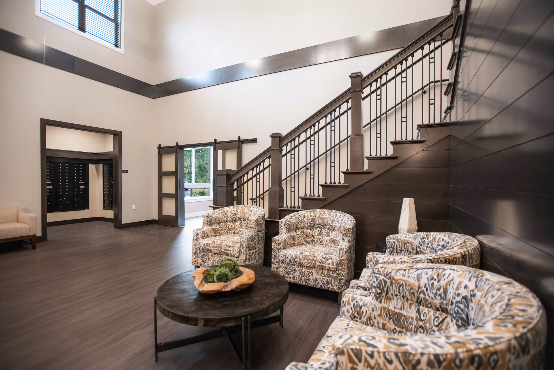 A spacious and modern interior common area with patterned armchairs arranged around a round wooden coffee table. There is a staircase with dark wood and metal railings on the right side, a large window letting in natural light, and a doorway leading to another room with mailboxes visible. The flooring is a dark wood tone and the walls are light-colored with dark trim accents.
