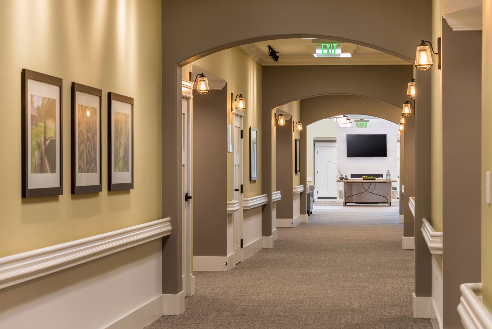 A well-lit hallway in a senior living facility with beige walls, framed artwork on the left wall, multiple wall-mounted light fixtures, and several doorways along the corridor. At the end of the hallway, there is a seating area with a flat-screen TV mounted above a console table.