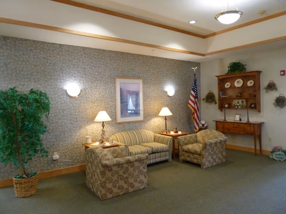 A cozy sitting area with a striped sofa, two patterned armchairs, side tables and lamps, an American flag, and wall decor in a common room.